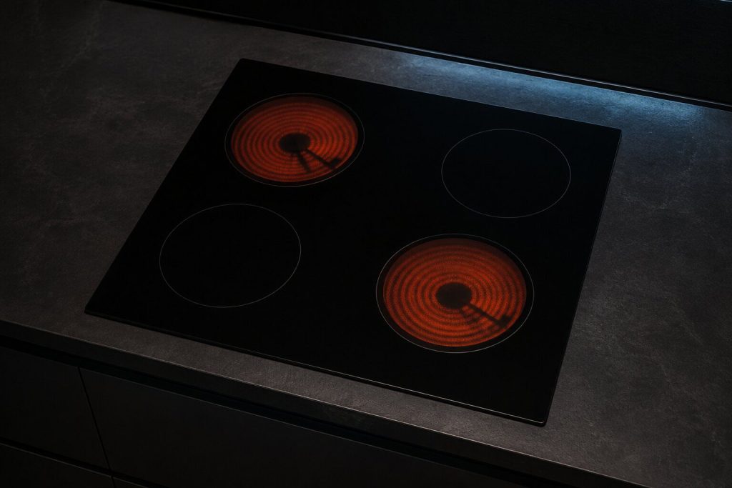 A smooth black ceramic cooktop set into a dark stone counter, photographed from above and slightly to the side in a near-black kitchen, two radiant coil zones glowing dull orange beneath the glass and the other two dark.