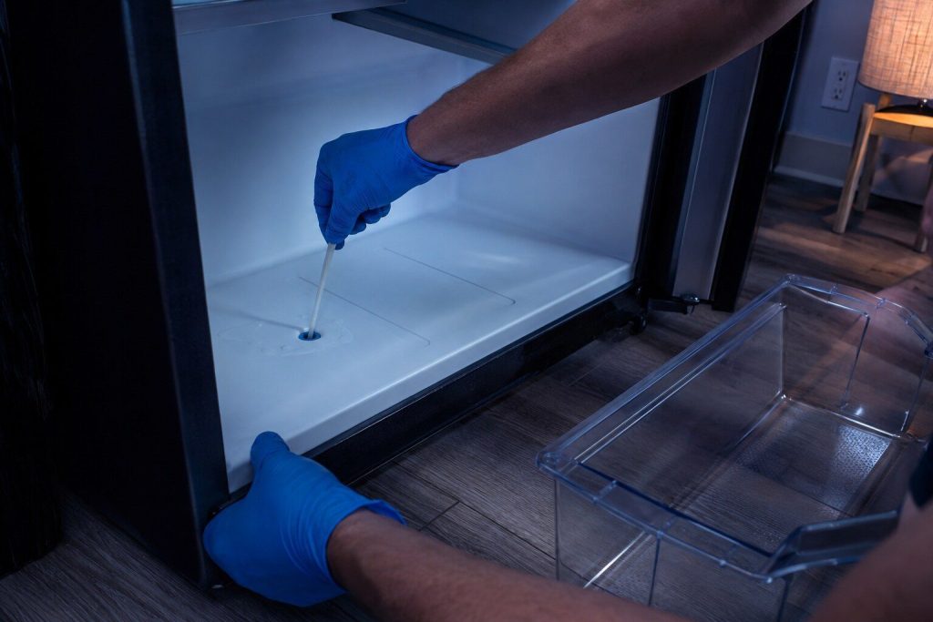 A crisper lifted out and a thin plastic blade clearing the small round drain hole at the back of the compartment floor. The exact object of the page, and plastic rather than wire, which is the safety point it makes.