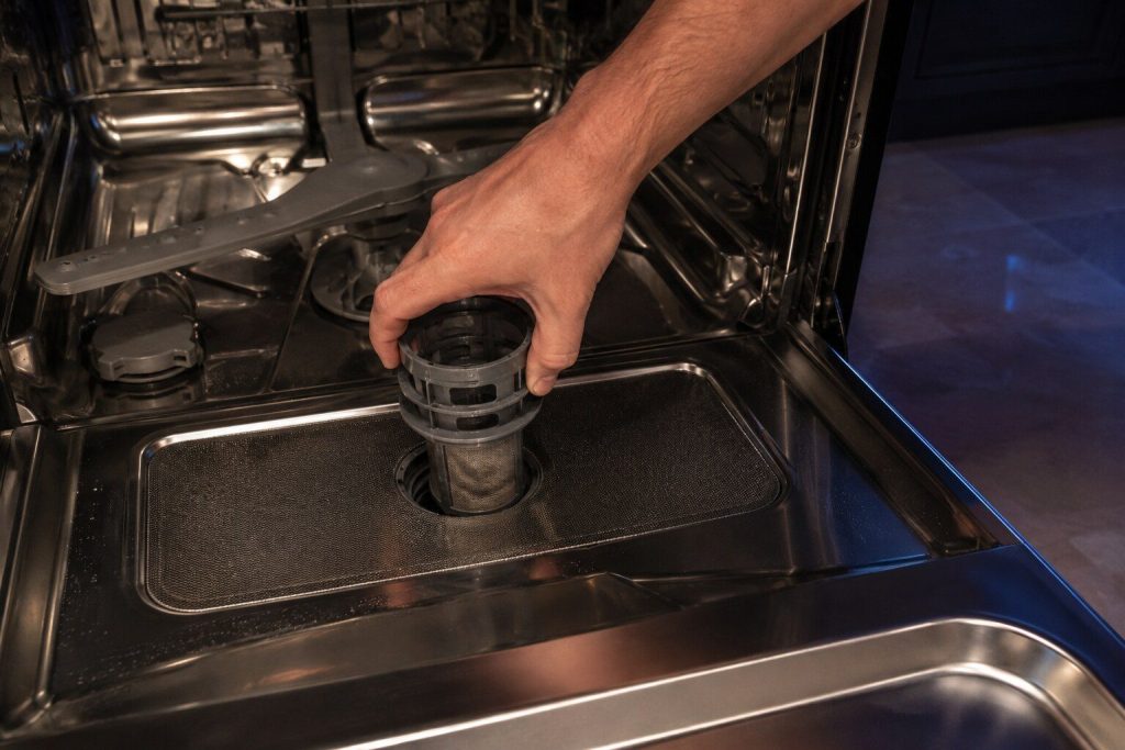 A hand turning the cylindrical filter counter-clockwise out of the floor of a dishwasher tub with the flat mesh plate still in place beneath it. Both filters in one frame, which is the detail most owners do not know about.