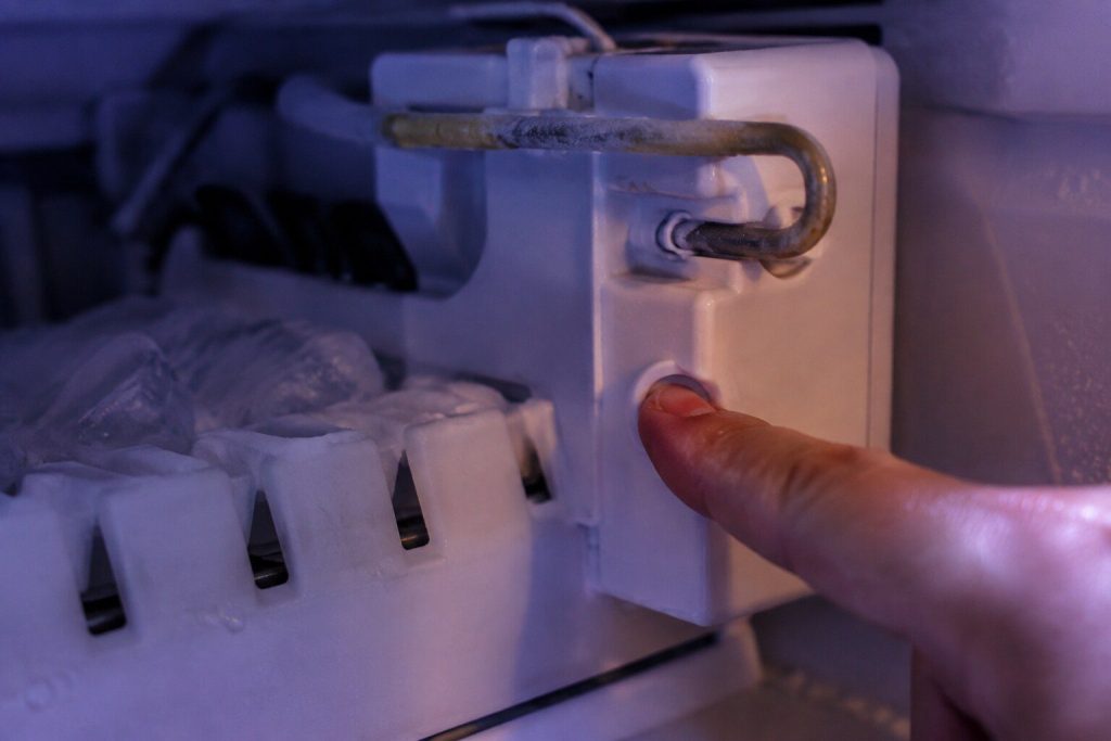 A finger pressed into the small recessed button on the side of an ice maker module inside a freezer compartment, the ejector arm above it. The button people search for, and the page explains what pressing it proves.