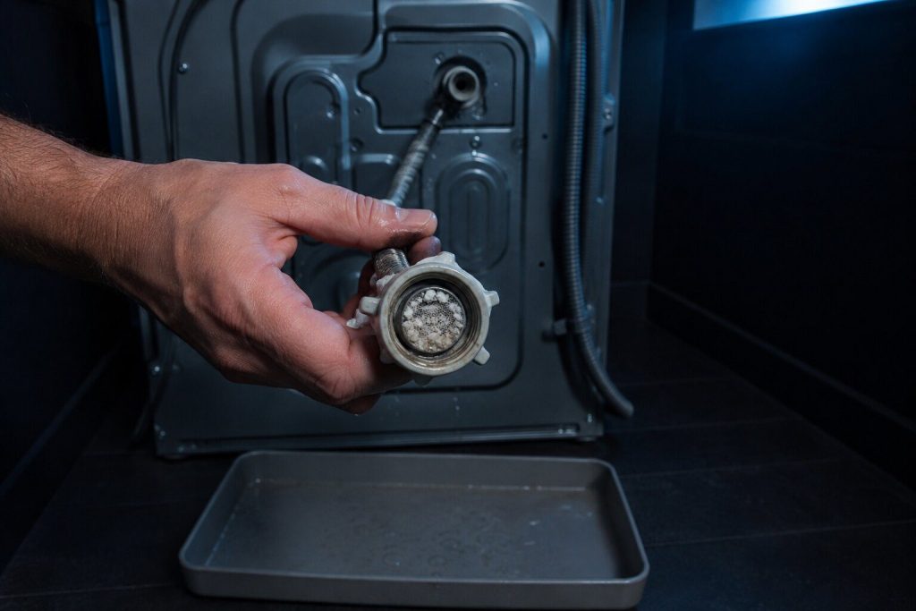 A braided supply hose unscrewed from the back of a washing machine and held so the small mesh screen inside its coupling faces the camera, the screen visibly furred with white scale, a shallow tray on the floor beneath.