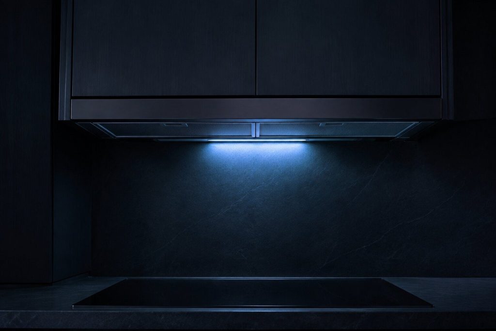 A slim under-cabinet range hood fitted beneath a wall cabinet above an empty cooktop, seen from below and slightly to the side, its mesh grease filters visible.