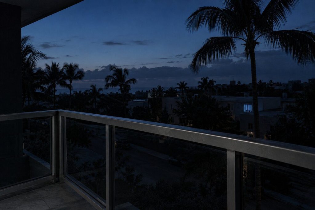 A condominium balcony rail at blue hour with a dark street and low buildings below, palm fronds against the last light