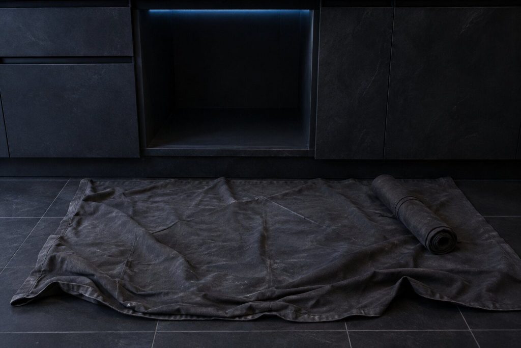 A canvas dust sheet spread across a near-black tile kitchen floor in front of an empty cabinet opening, a rolled tool mat lying closed at one edge