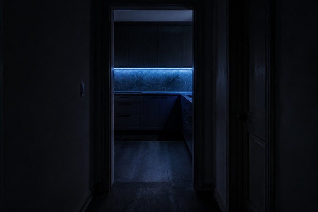 A dark hallway looking through a doorway into a kitchen lit only by one under-cabinet strip of ice-blue light, nobody in either room
