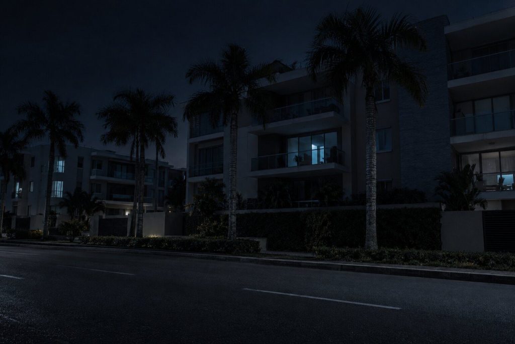 A quiet residential street of low condominium blocks at night seen from across the road, palms along the pavement and a few cold lit windows