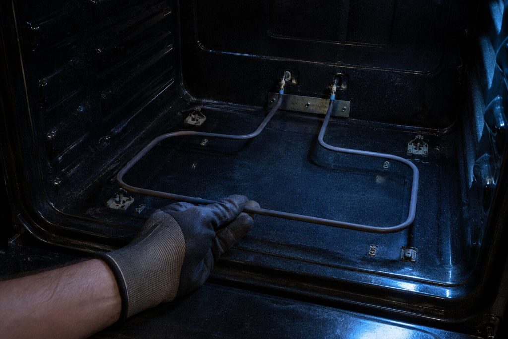 A bake element unbolted from the floor of an oven cavity and drawn forward a hand's width on its two leads, held in a gloved hand, the cavity dark behind and one work light raking in from the open door.