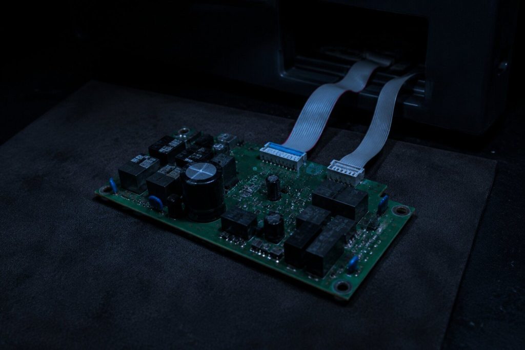 A green control board lifted out of an appliance and laid on a dark antistatic mat under one low lamp, two ribbon connectors still trailing back into the opening, relays and a large capacitor visible, nothing on the board readable.