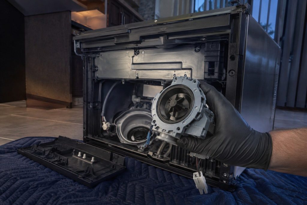 A dishwasher on its back with the base cover off and the drain pump lifted clear of the sump on a gloved hand, the impeller housing turned to the camera. The impeller is where the glass ends up, which is the story this page tells.