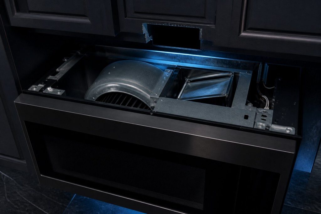 The top cover removed from an over-the-range microwave still on its mounting, the squirrel cage blower and its vent flap visible inside from above with the flap turned to the other setting, the duct opening in the cabinet above it.
