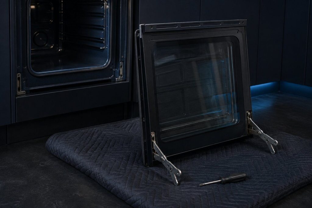 An oven door lifted off a range and stood upright against a padded blanket on a near-black floor, both hinge arms locked open and visible, the empty hinge slots in the oven frame behind it, a screwdriver lying on the blanket.