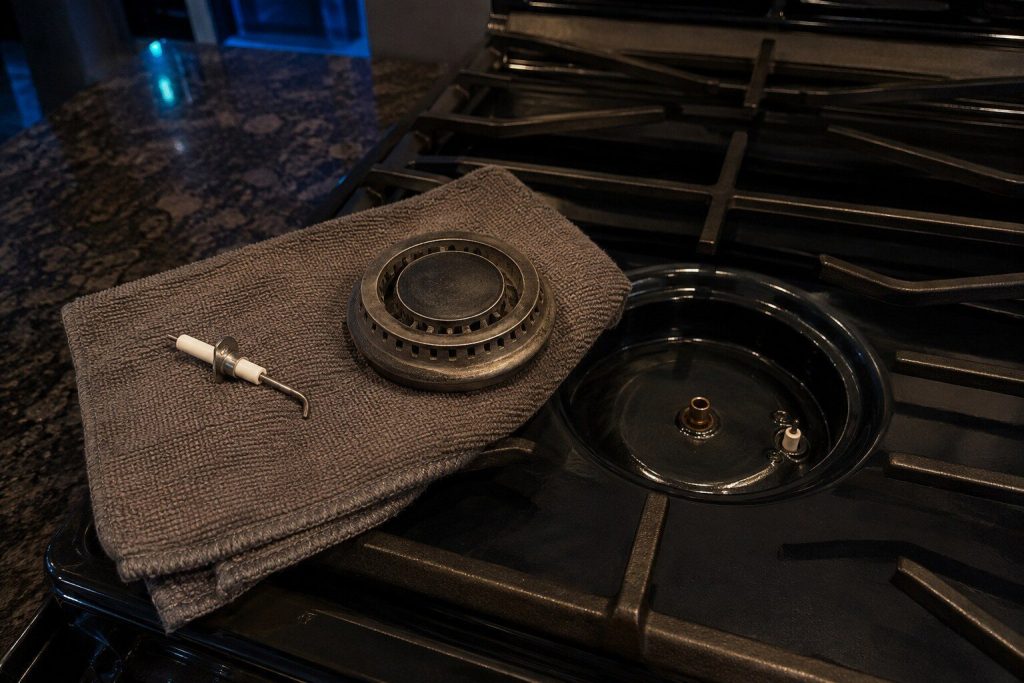 A spark igniter and a burner head lying apart on a folded towel on a cooktop, the empty burner base and its orifice beside them. The endless click is one of these two and the frame shows both.