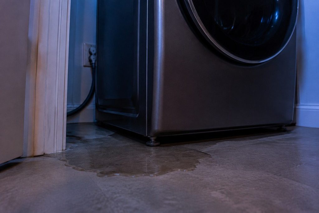 A dark wet patch spreading from beneath the front left corner of a washer with the leveling foot standing in it. Where the water ends up is not where it left, which is the sentence this page opens with.