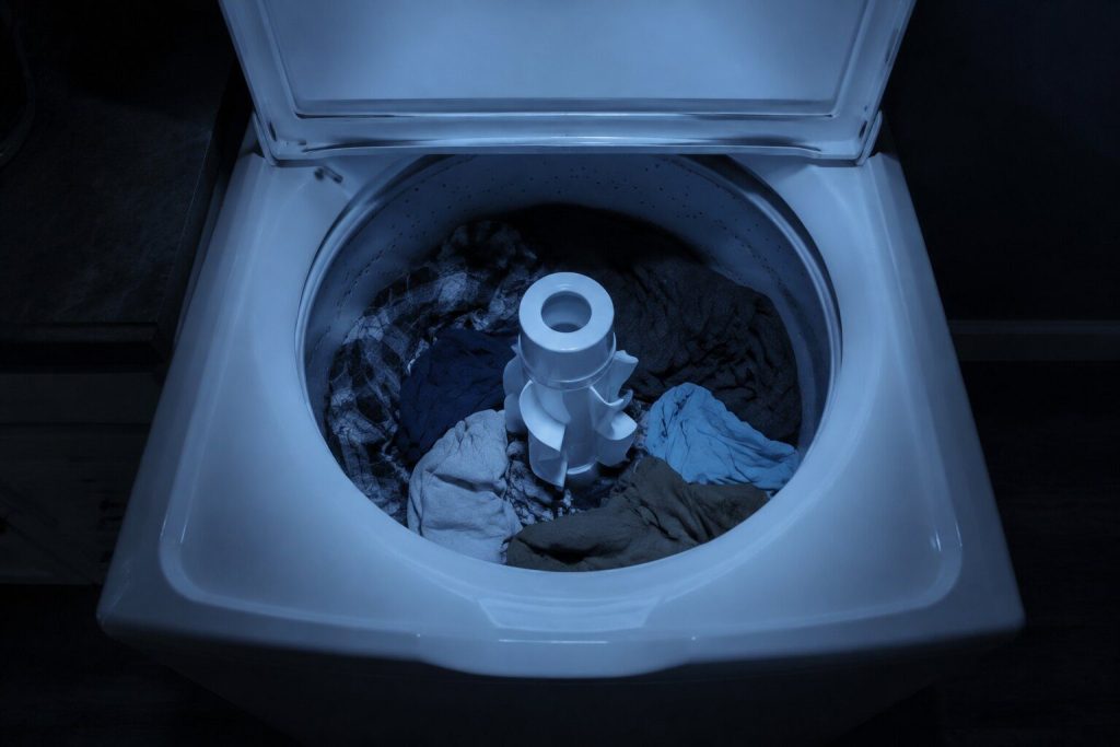 Looking down into the open tub of a white top-load washer at night: a load of laundry lying loose and completely dry around a tall finned center agitator post, not a drop of water anywhere in the basket.