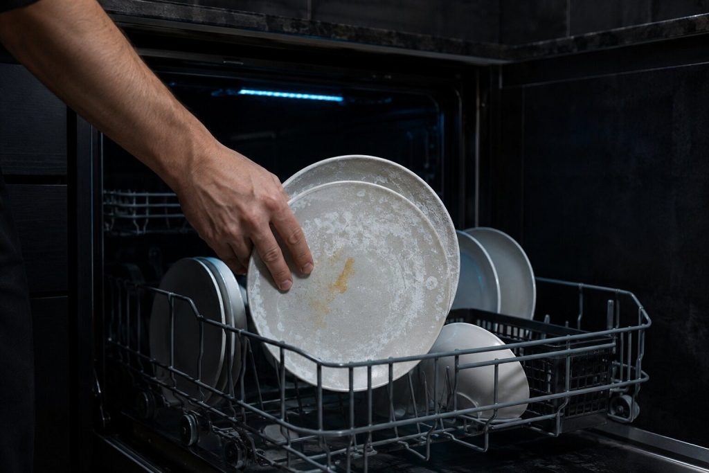 A hand lifting two plates from the lower rack of an open dishwasher in a near-black kitchen, both carrying a chalky white film and a smear of dried food, the rack behind them still loaded and the tub dark.