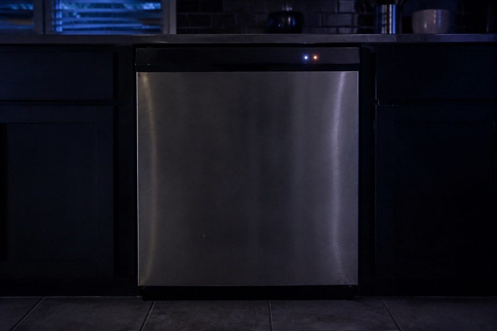 A dishwasher front in a dark kitchen with two small round indicator lamps lit on its top edge and everything else black